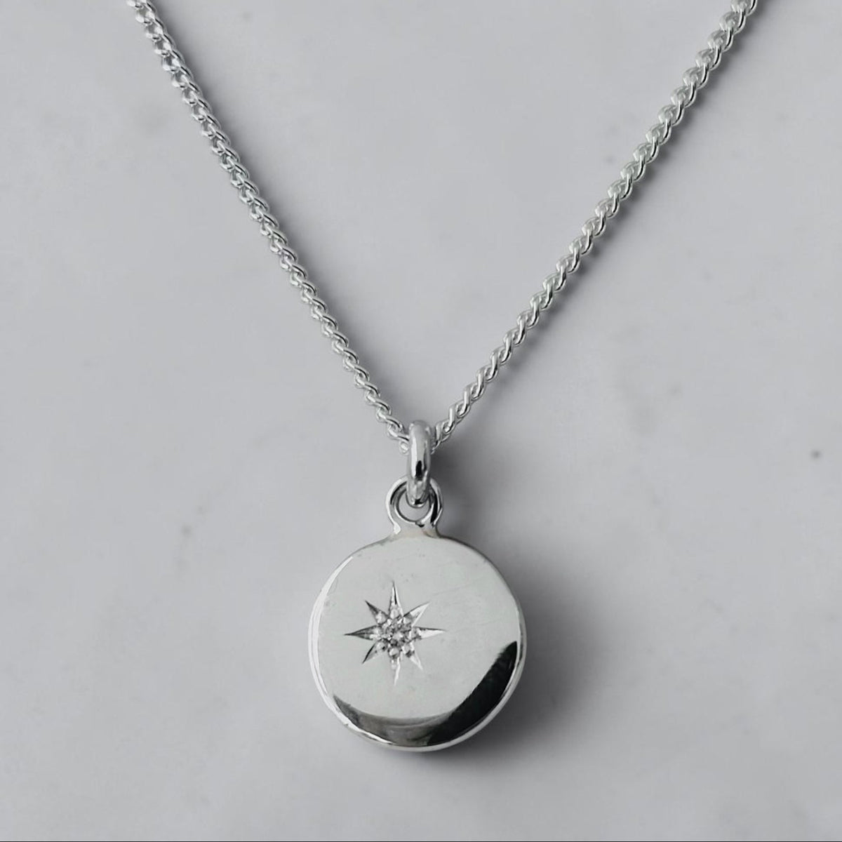 A silver chain necklace with a round disc pendant featuring a small starburst set diamond design, photographed on a light gray marble surface.