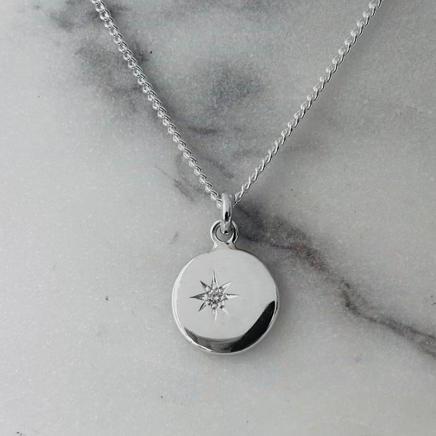 A silver round disc pendant necklace with a small starburst design and a clear central diamond, displayed on a light gray marble surface.