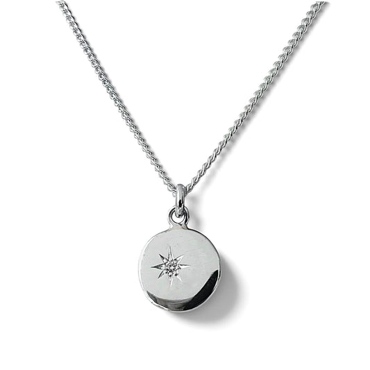 A silver chain necklace with a round disc pendant featuring an engraved star design and a small diamond set in a starburst design, shown on a white background.